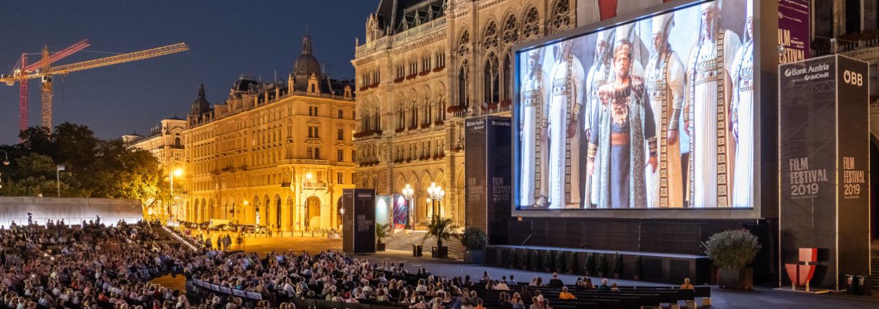 Auf einer am Wiener Rathaus befestigten Leinwand wird vor zahlreichen sitzenden Zuschauer*innen ein Opernfilm gezeigt.