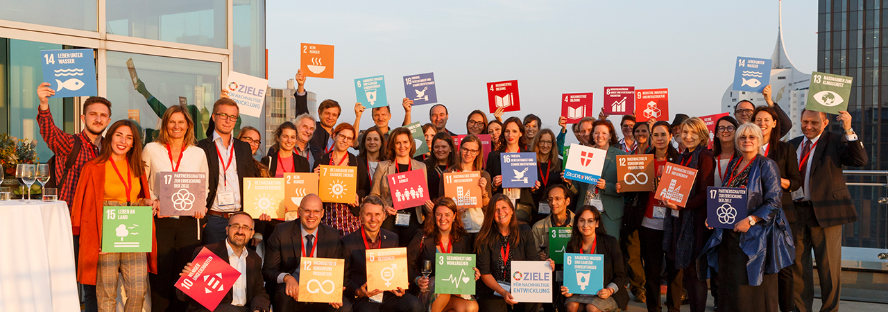 Participants of the EC-Conference 'Sustainable Development Goals' are standing on a terrace are holding up the 17 UN Development Goals on colorful cards