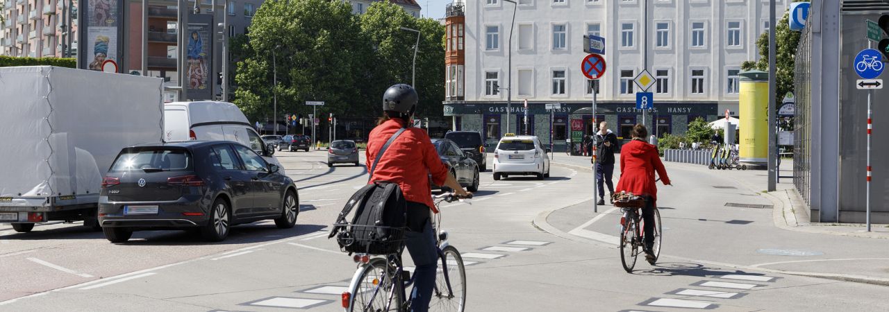 Fahrradfahrerinnen mit roten Jacken überfahren eine Straße auf einem Radfahrübergang, neben ihnen fahren verschiedene Autos