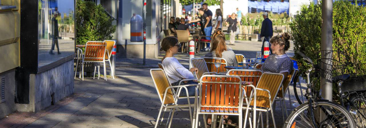 Drei Frauen sitzen auf orangen Gartenstühlen auf einem Platz vor einem Lokal.