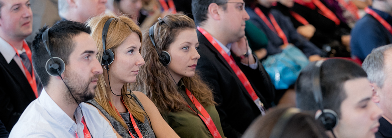 Teilnehmer*innen mit roten Lanyards und Dolmetsch-Kopfhörern blicken konzentriert in Richtung Vortrag