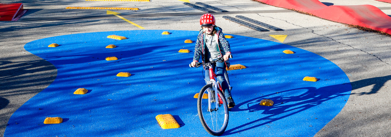 Kind auf rotem Fahrrad und mit rotem Fahrradhelm fährt auf blauem Untergrund und gelben Hindernissen.