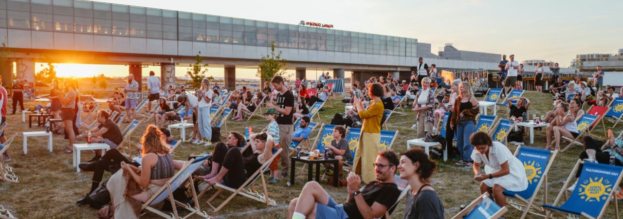 Menschen bei Sonnenuntergang in Liegestühlen auf einer Wiese, im Hintergrund die U-Bahn-Station Seestadt
