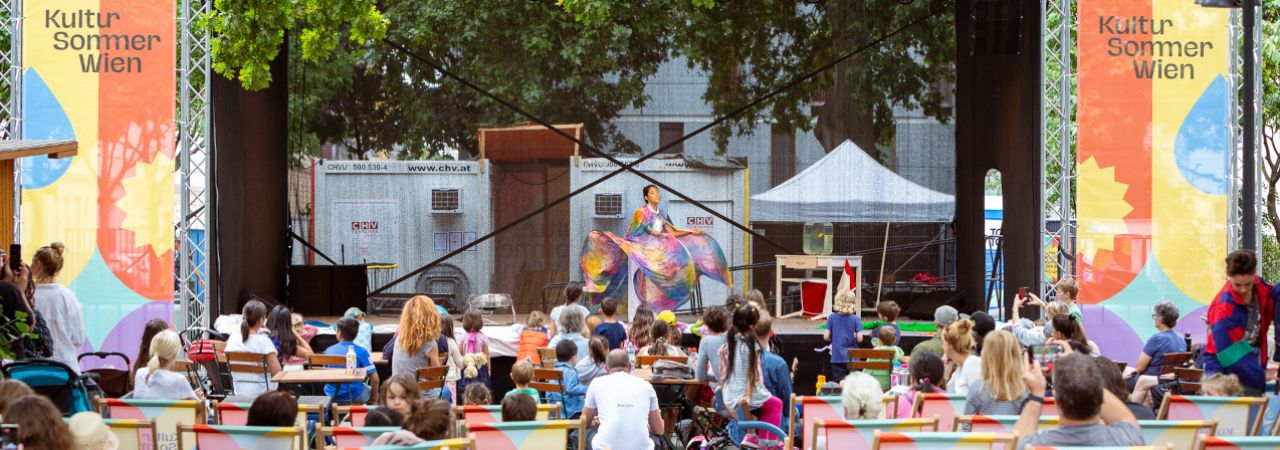 Open-Air-Bühne mit Künstlerin, davor Zuschauer*innen in Liegestühlen