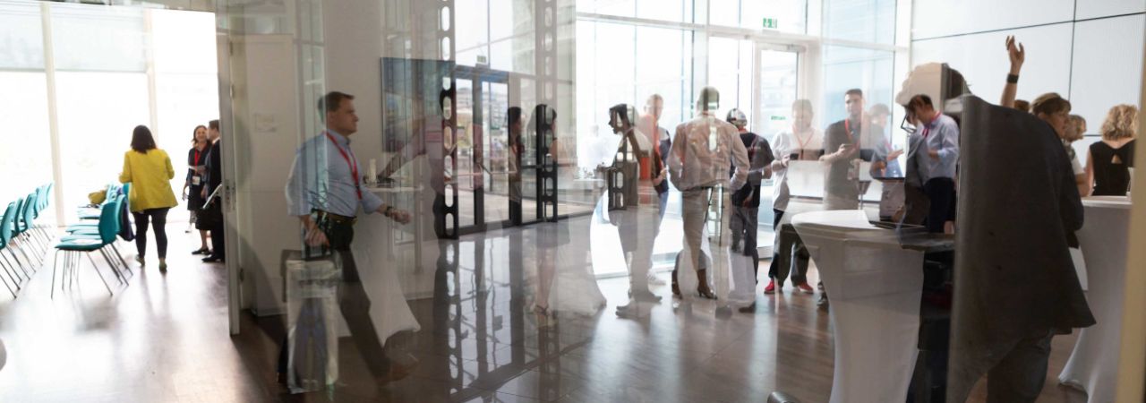 Konferenzteilnehmer*innen an Stehtischen im Gespräch, hinter einer Glasscheibe fotografiert.