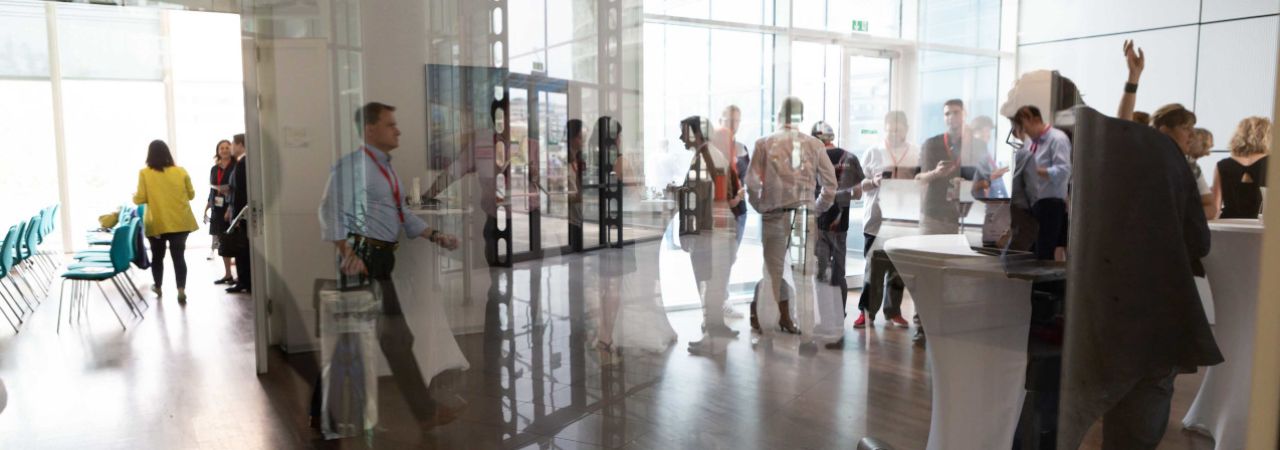 Personen bei einer Konferenz an Stehtischen, fotografiert durch eine Glasscheibe