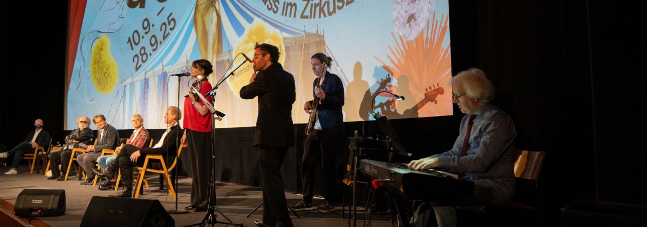 Musiker*innen mit Instrumenten auf der Bühne vor dem Produktions-Sujet.