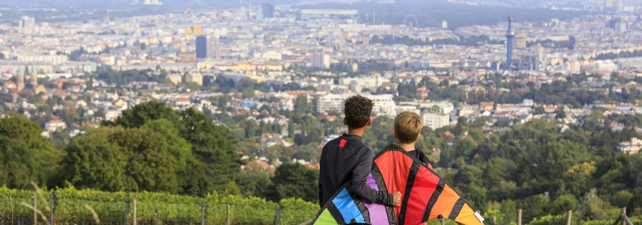 Zwei Kinder mit Flugdrachen schauen von Berg auf Wien hinunter