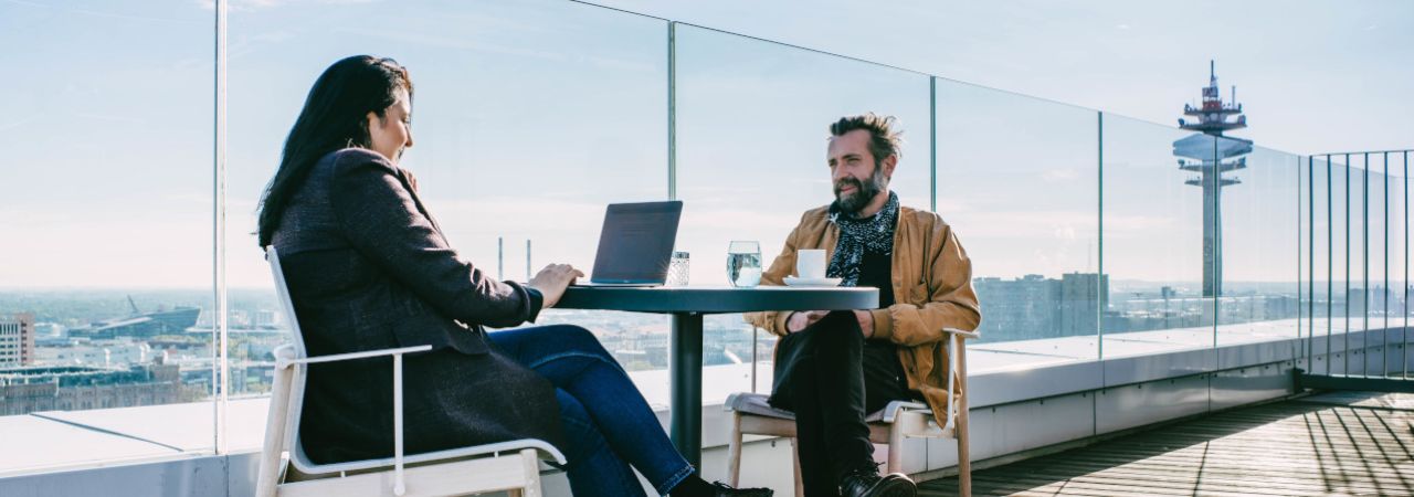 Eine Frau und ein Mann sitzen auf einer Dachterrasse an einem Kaffeehaustisch. Die Frau arbeitet auf einem Laptop.