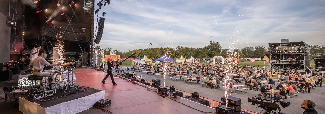 Open-Air-Konzert auf einer großen Bühne, davor ZuschauerInnen auf Heurigengarnituren